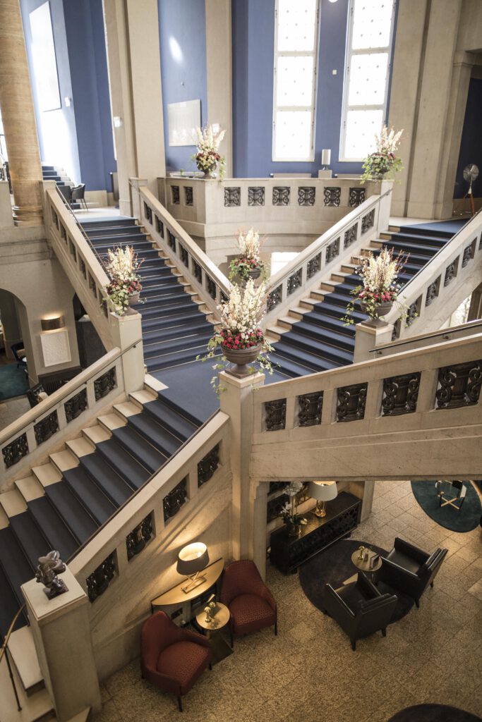 Aufwendig dekorierte historische Treppenanlage in der Eingangshalle des Hotel The Wellem Düsseldorf mit üppigen Blumenarrangements, blauen Wandakzenten und gemütlichen Loungebereichen im Untergeschoss.