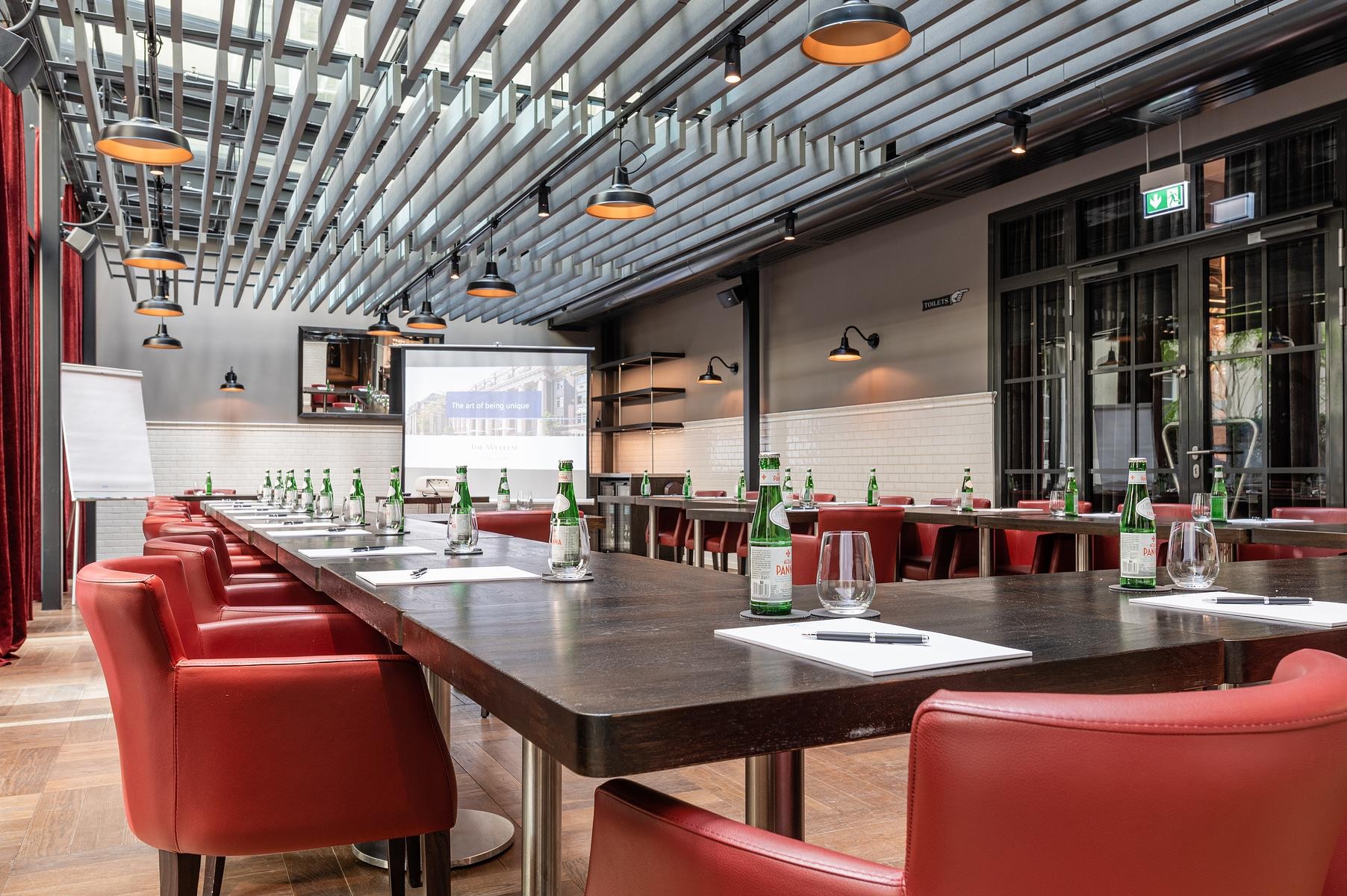 Wintergarten Eventfläche im Hotel The Wellem in Düsseldorf mit U-förmiger Bestuhlung, roten Ledersesseln, großem Tagungstisch, Präsentationsleinwand und modernem Oberlicht für lichtdurchflutete Meetings.
