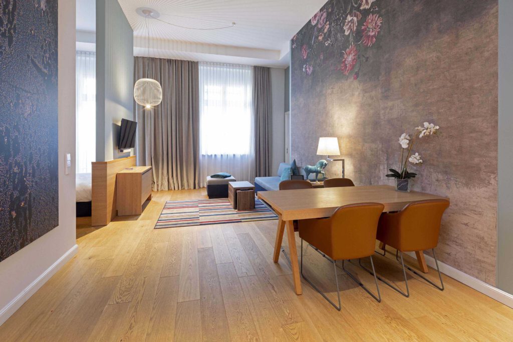 Elegante Executive Suite im Hotel The Wellem Düsseldorf mit großem Wohnbereich, Essplatz aus Holz, stilvoller Blumenakzentwand, Designsofa und separatem Schlafbereich.
