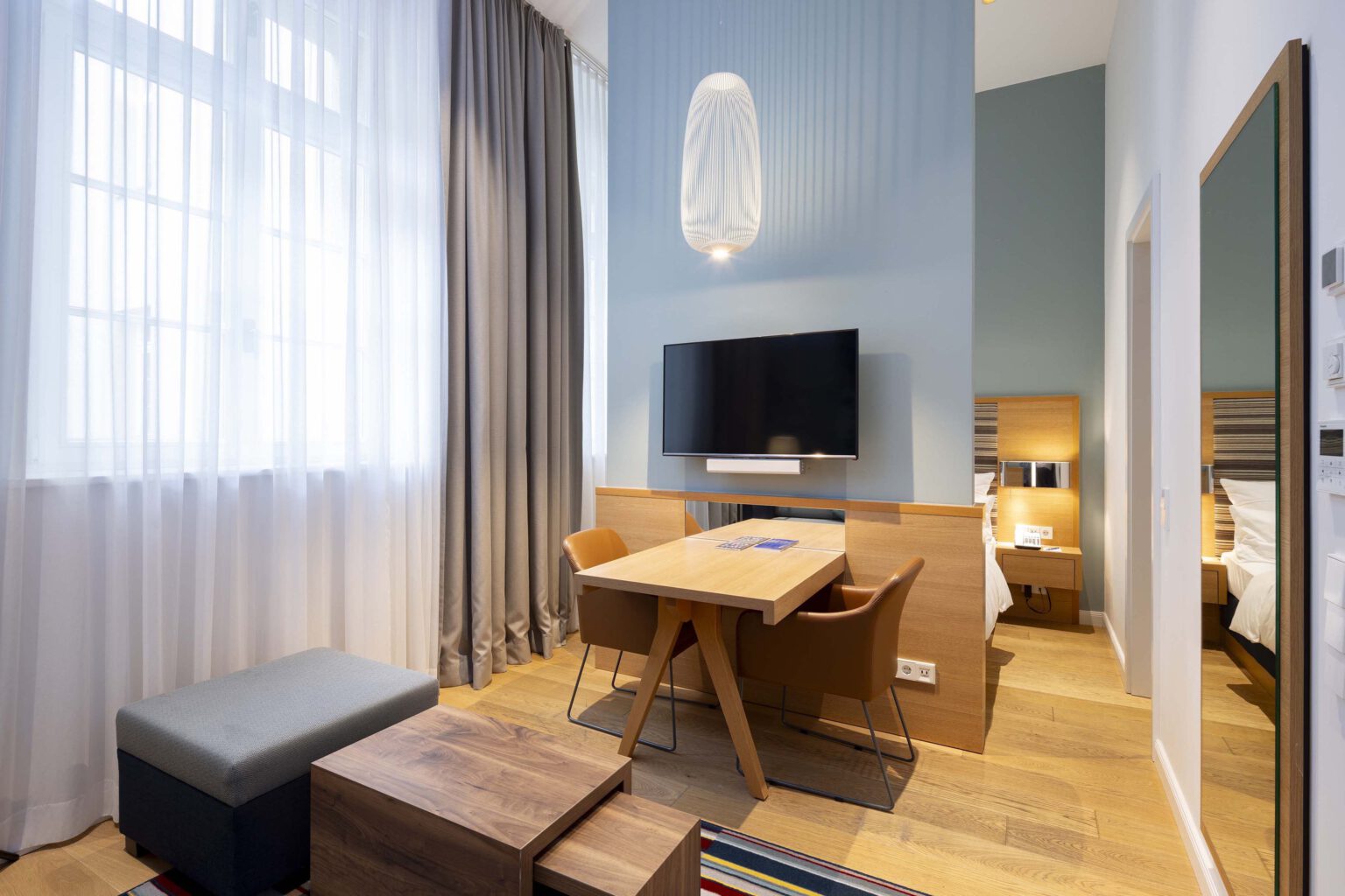 Modernes Studio im Hotel The Wellem Düsseldorf mit Schreibtisch, TV, warmen Holzakzenten und Blick in den hellen Schlafbereich mit komfortablem Doppelbett.