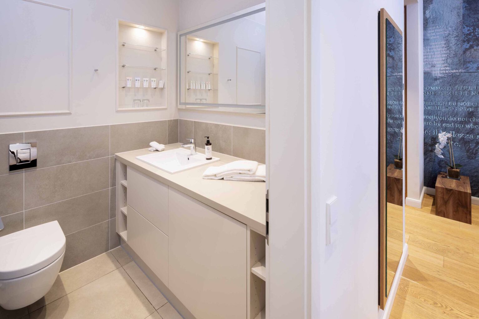 Modern bathroom in the studio room at The Wellem Hotel Düsseldorf with large washbasin, illuminated mirror, open shelves and elegant tiles in beige and gray tones.