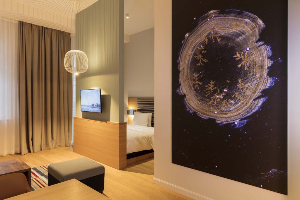 Helle Studio Suite mit modernem Wohn- und Schlafbereich, großem abstrakten Kunstwerk, elegantem Holzinterieur und gemütlicher Beleuchtung im Hotel The Wellem in Düsseldorf.