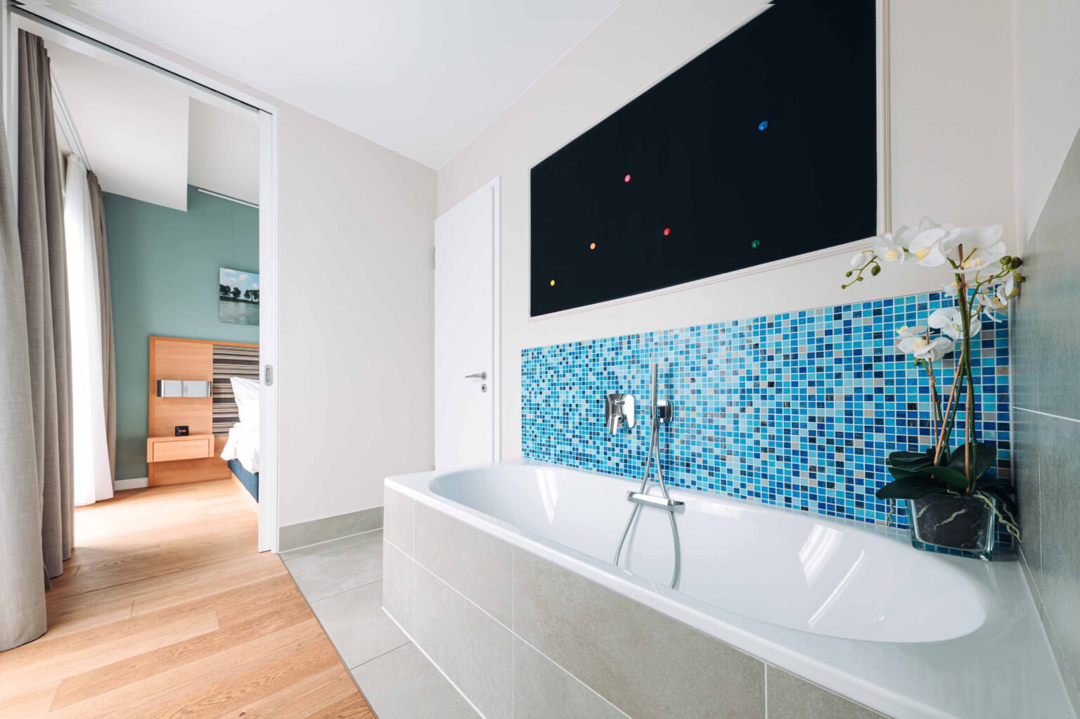 Modern bathroom in the Studio Suite with elegant bathtub, blue mosaic tiles, discreet wall art and view into the bright bedroom.