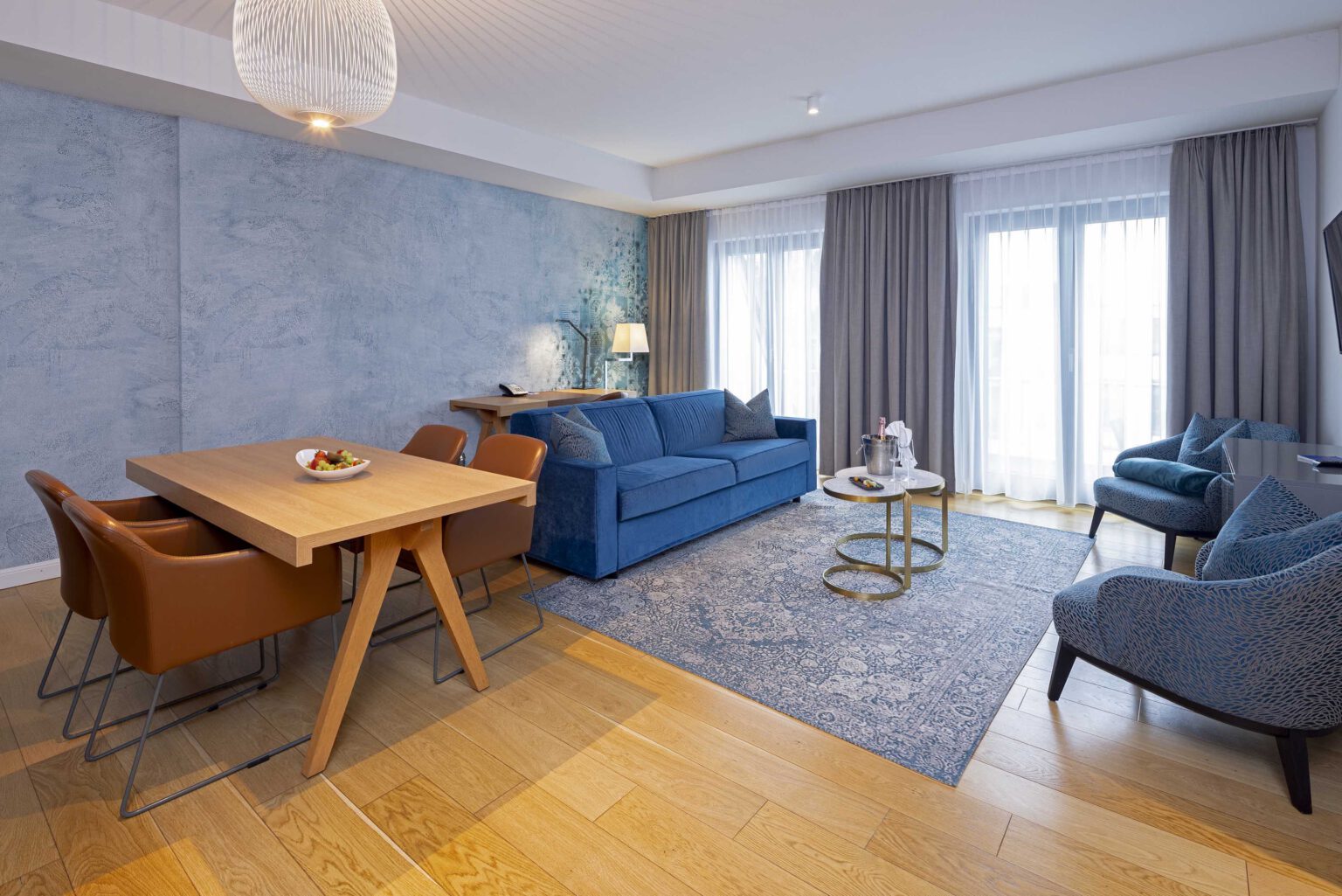 Großzügiger Wohnbereich der The Wellem Suite mit elegantem Holztisch, samtblauer Couch, designstarken Sesseln, bodentiefen Fenstern und stilvoller Akzentwand im luxuriösen Hotel The Wellem in Düsseldorf.