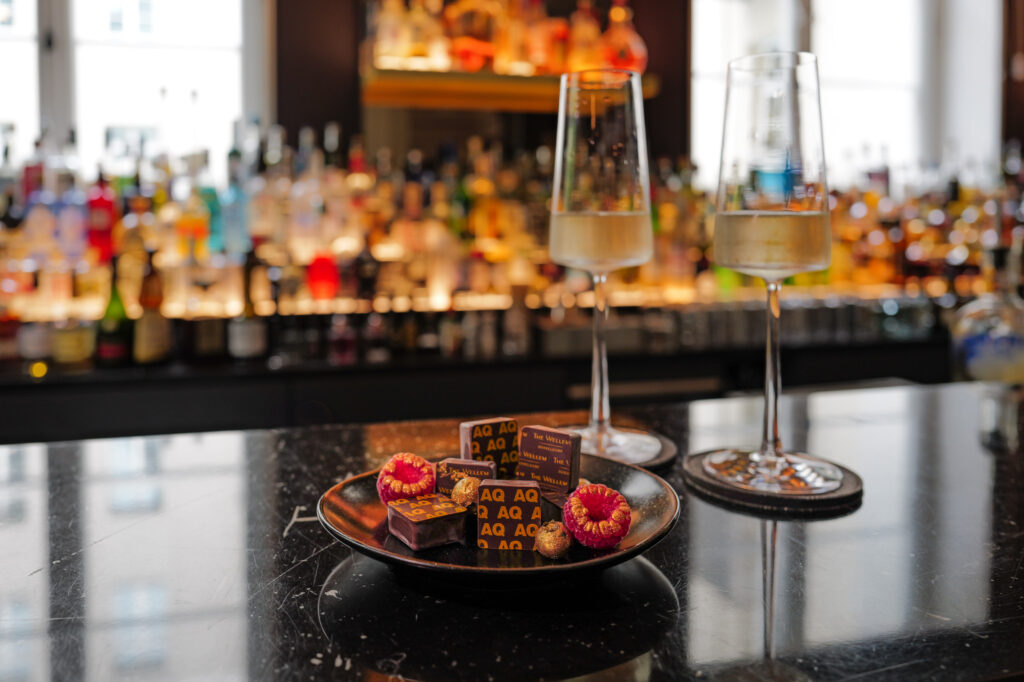Edle Pralinen mit AQ-Branding und frischen Himbeeren neben zwei Gläsern Sekt auf der The Wellem Bar im Hotel The Wellem Düsseldorf.