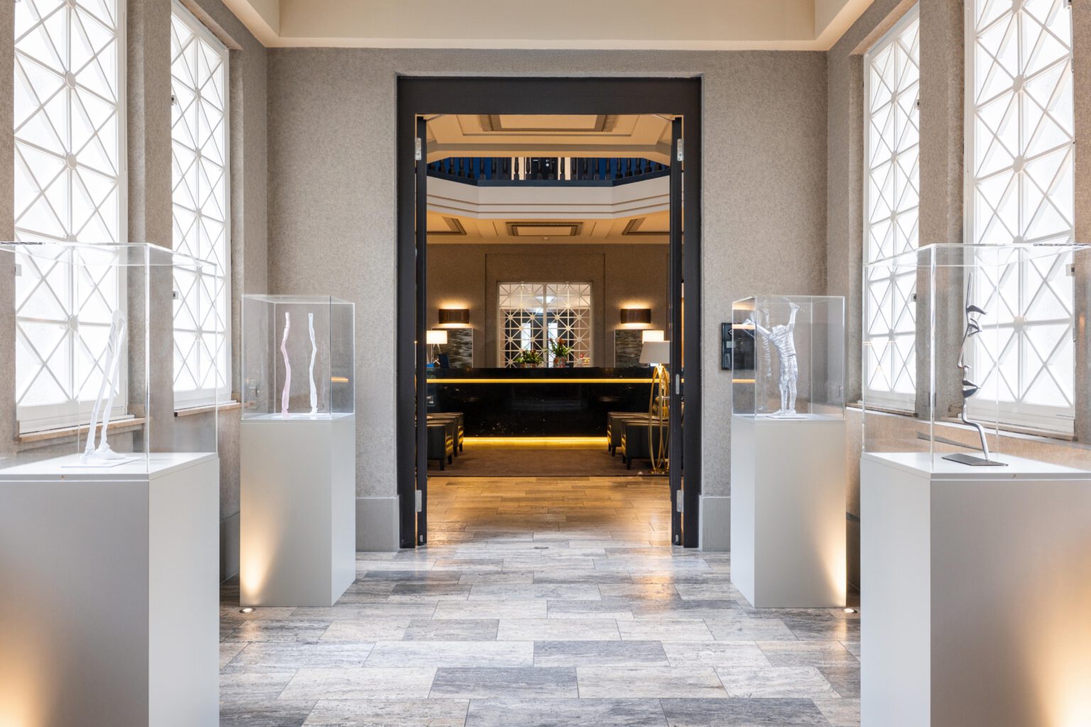 Bright art corridor at The Wellem Hotel in Düsseldorf with modern sculptures in glass cubes, leading directly into the elegant concierge lobby with marble counter and stylish seating areas.