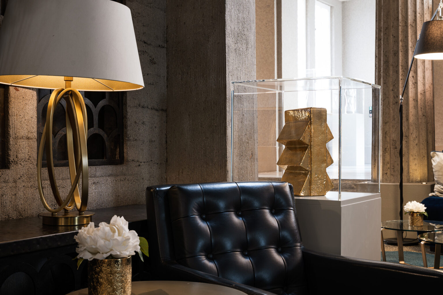 Elegant lounge at Hotel The Wellem Düsseldorf with modern table lamp, black leather armchairs and gold-colored art sculpture in a transparent display case in front of historic stone architecture.