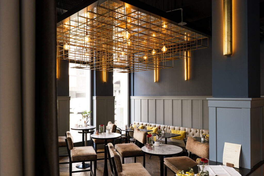 Modernes Interieur des Restaurants Le Cou Cou im The Wellem Düsseldorf mit goldener Raster-Deckeninstallation, runden Marmortischen und gemütlichen Sitzbereichen.
