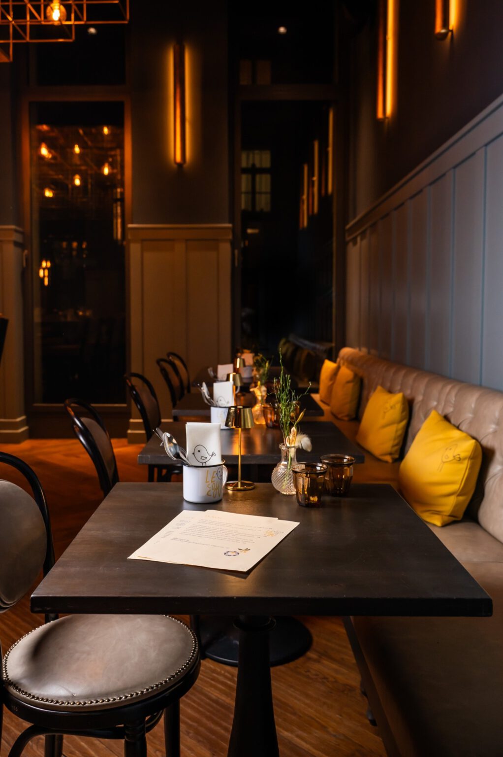 Eleganter Innenbereich des Restaurants Le Cou Cou in Düsseldorf mit dunklen Tischen, gedämpfter Beleuchtung, gelben Kissen und liebevoll gedeckten Plätzen.